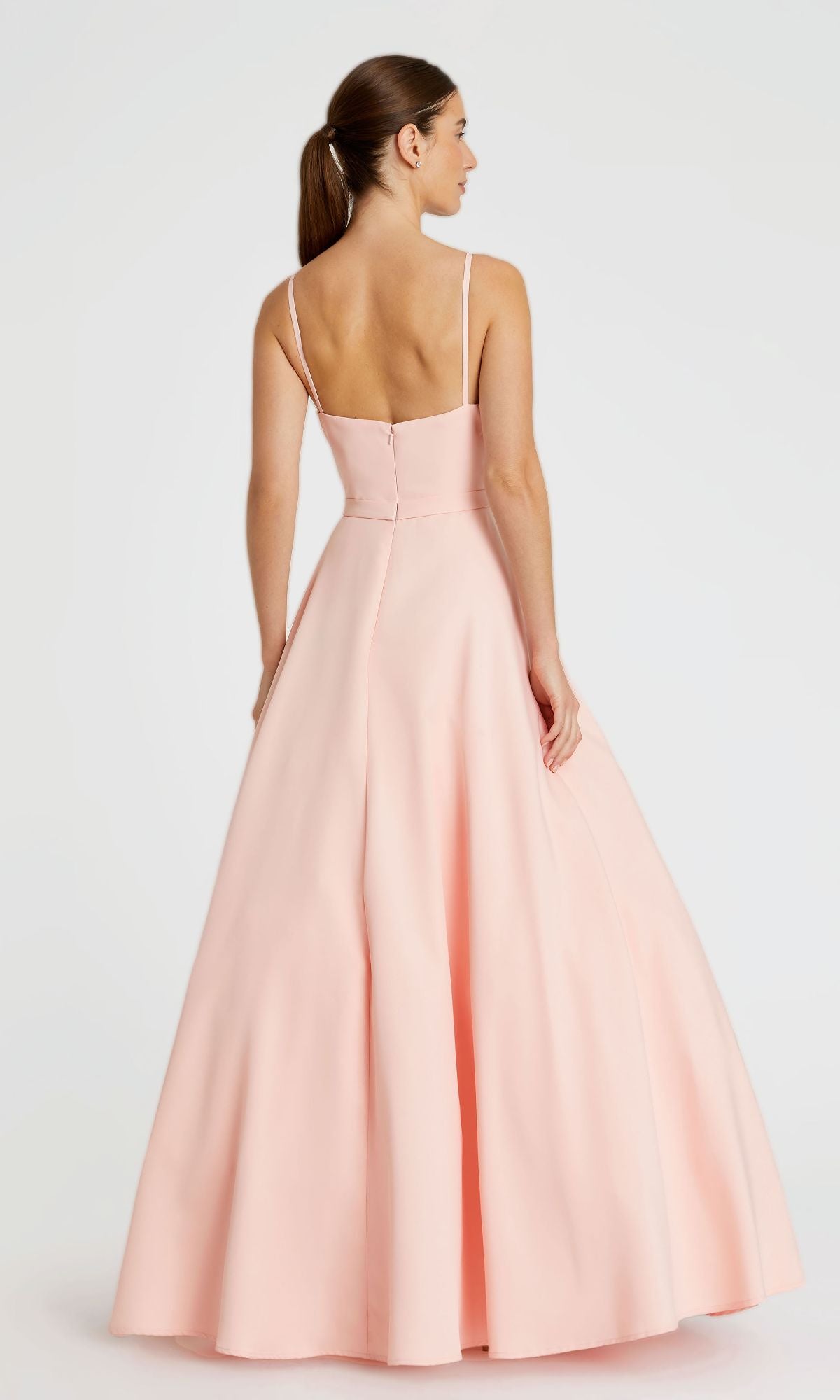 A woman stands facing away, dressed in the Classic Long A-Line Formal Dress: Mac Duggal 48855, a floor-length light pink gown featuring thin straps and a flared skirt.